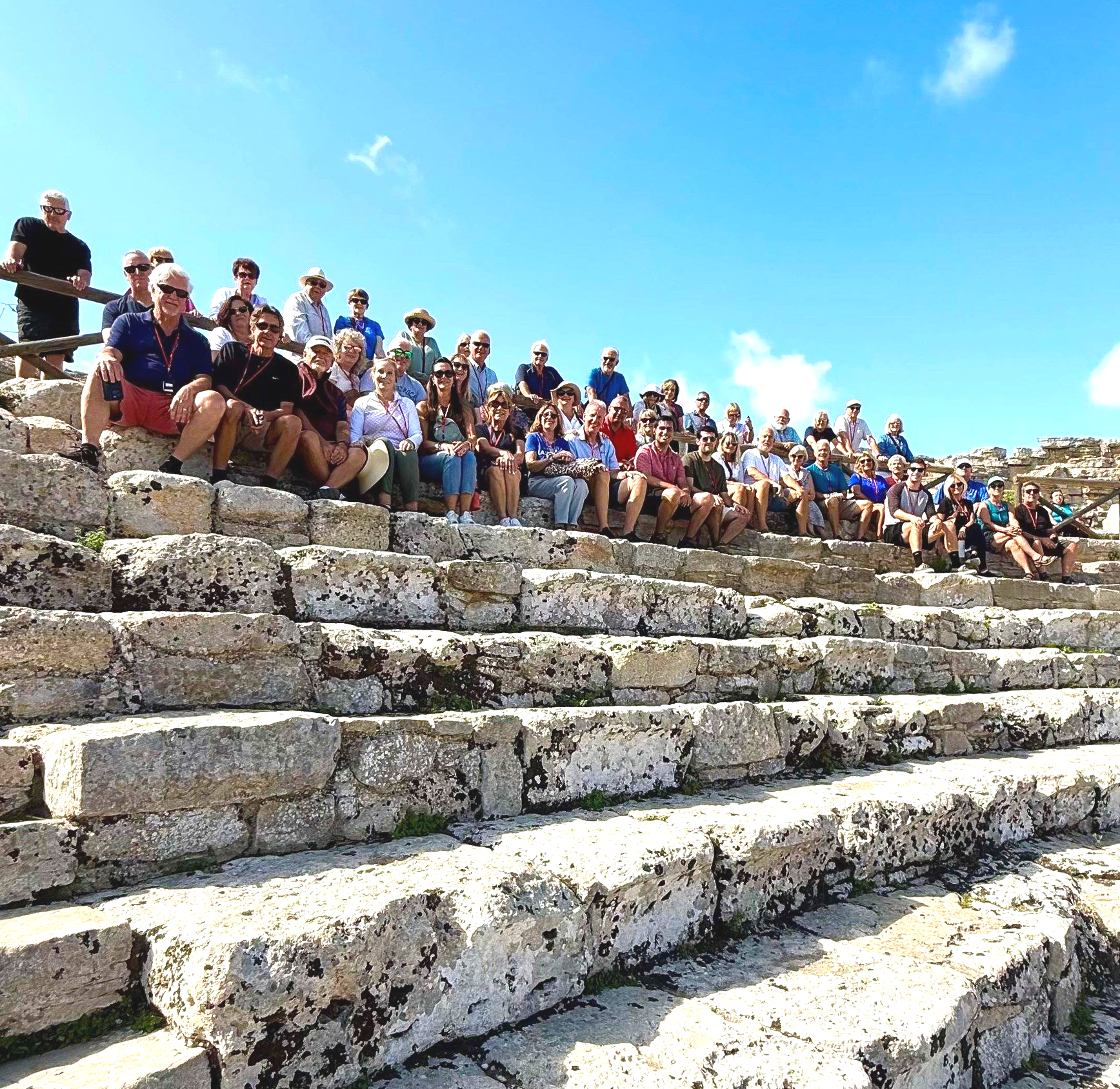 Group of people seated on stone steps under a bright blue sky.