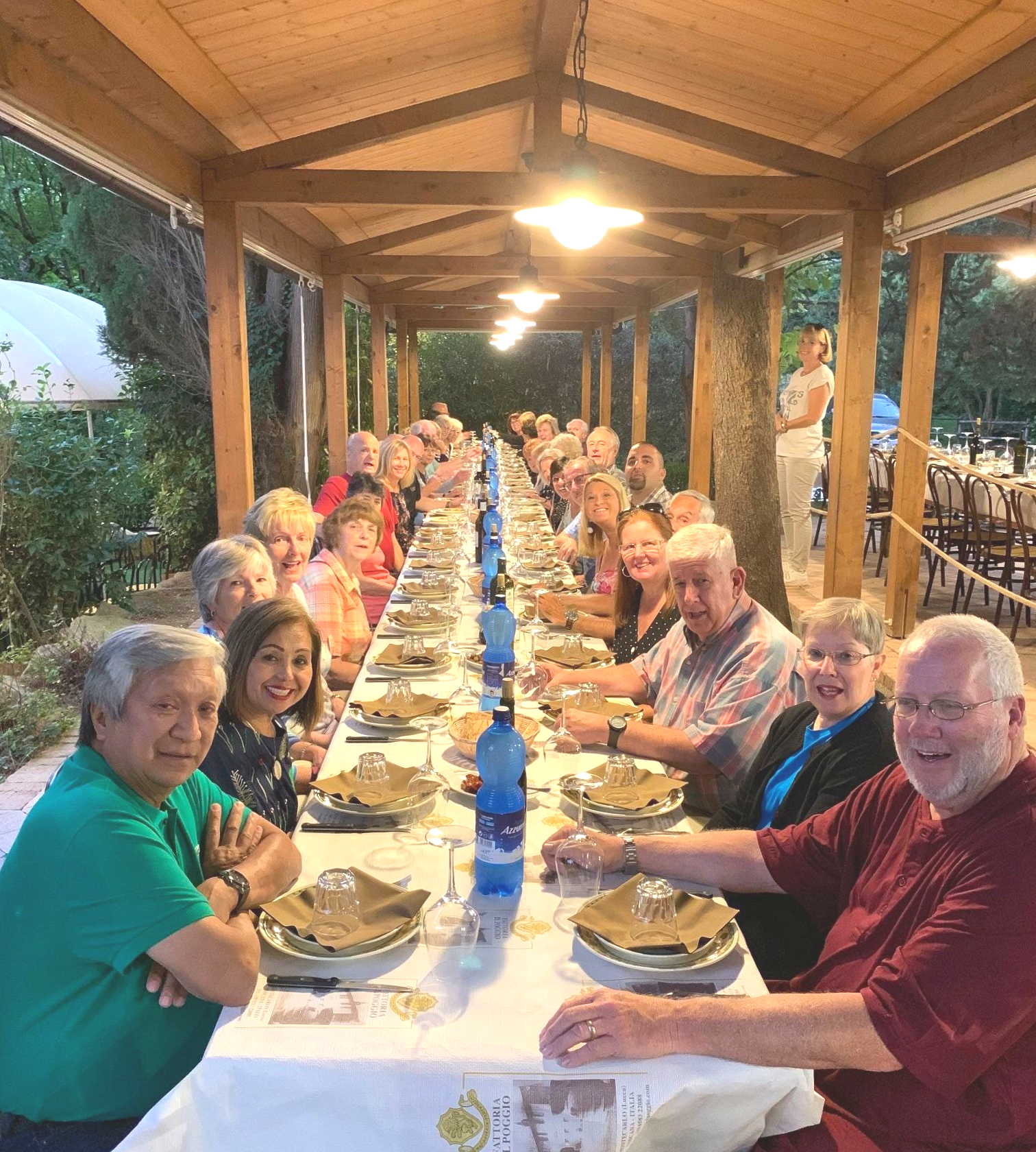 People seated at long table under a wooden pergola, dining outdoors.