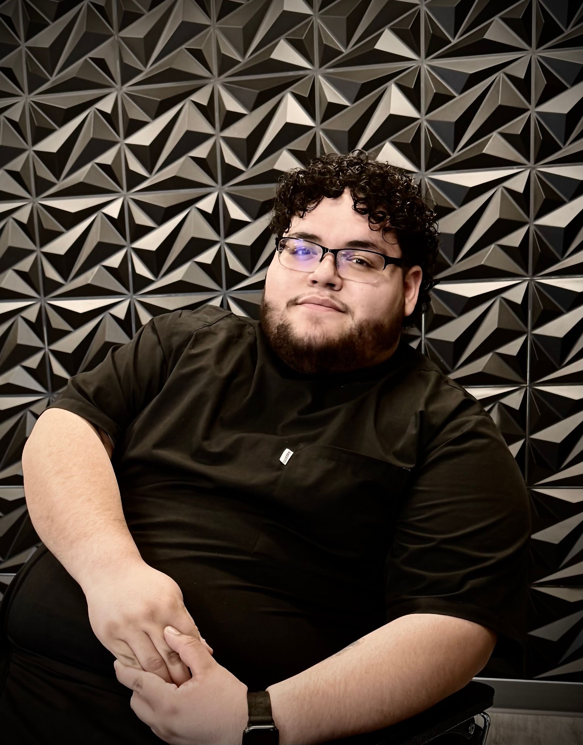 Man wearing glasses and black shirt, sitting in front of a geometric black wall.
