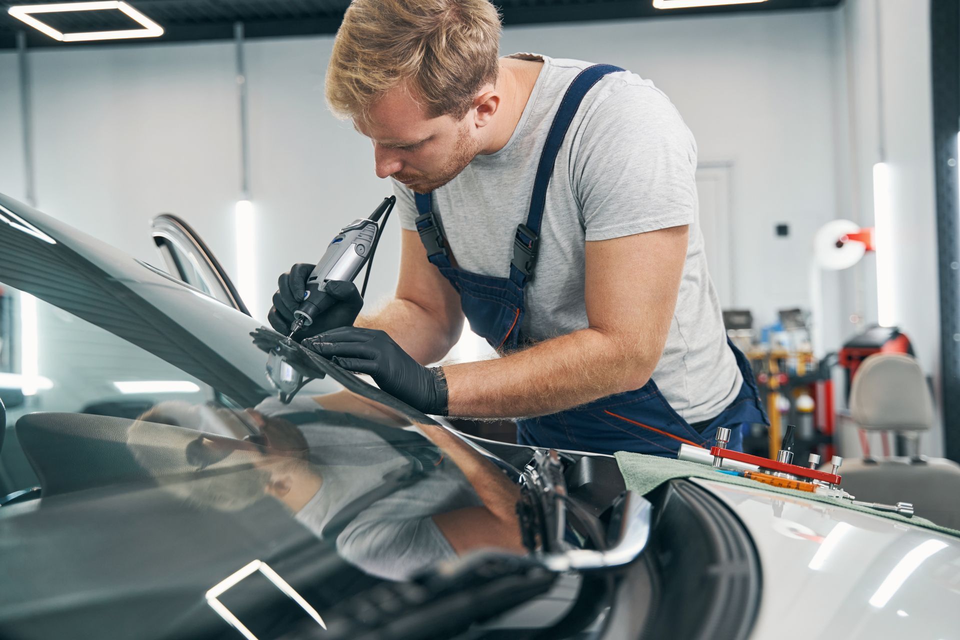 Mechanic in overalls, fixing a car windshield with a tool in a shop.