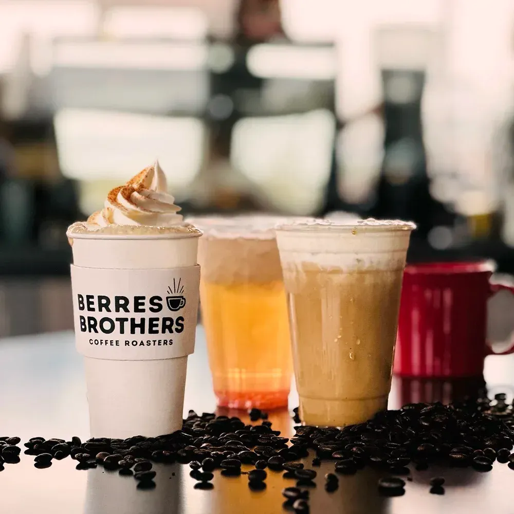 Four beverages, including a whipped cream-topped coffee, sit on a counter with scattered coffee beans.