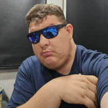 Person wearing blue sunglasses and a navy shirt leans on a table against a dark background.