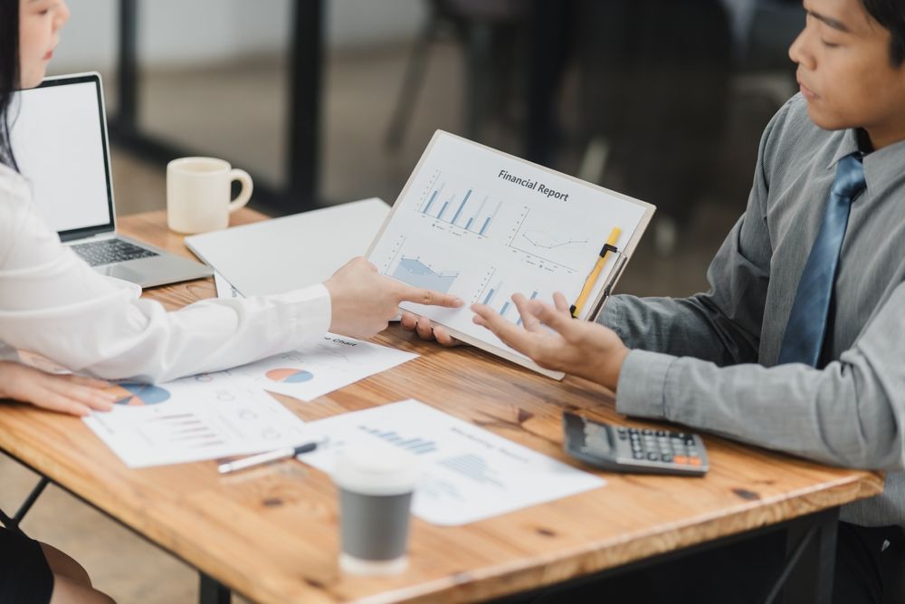 Two people discussing printed charts at a conference table with a laptop and coffee cup