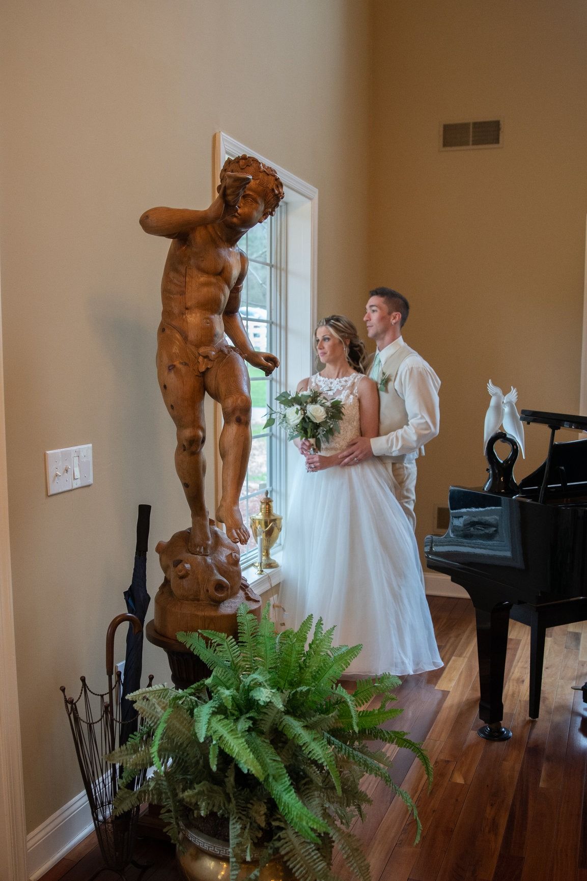 Wedding couple poses by sculpture and piano; woman in white dress, man in tan suit, looking at each other.