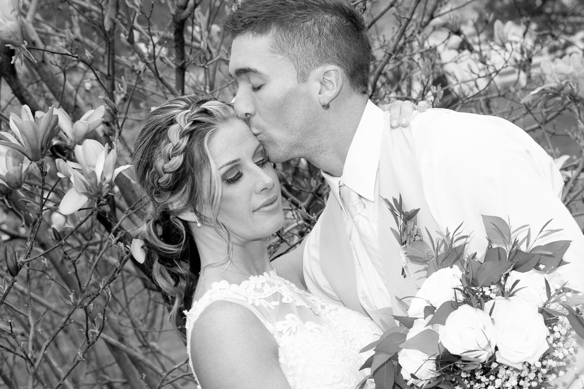 Groom kisses bride's forehead in wedding photo. Bride in lace dress, holds bouquet. Outdoors, black and white.