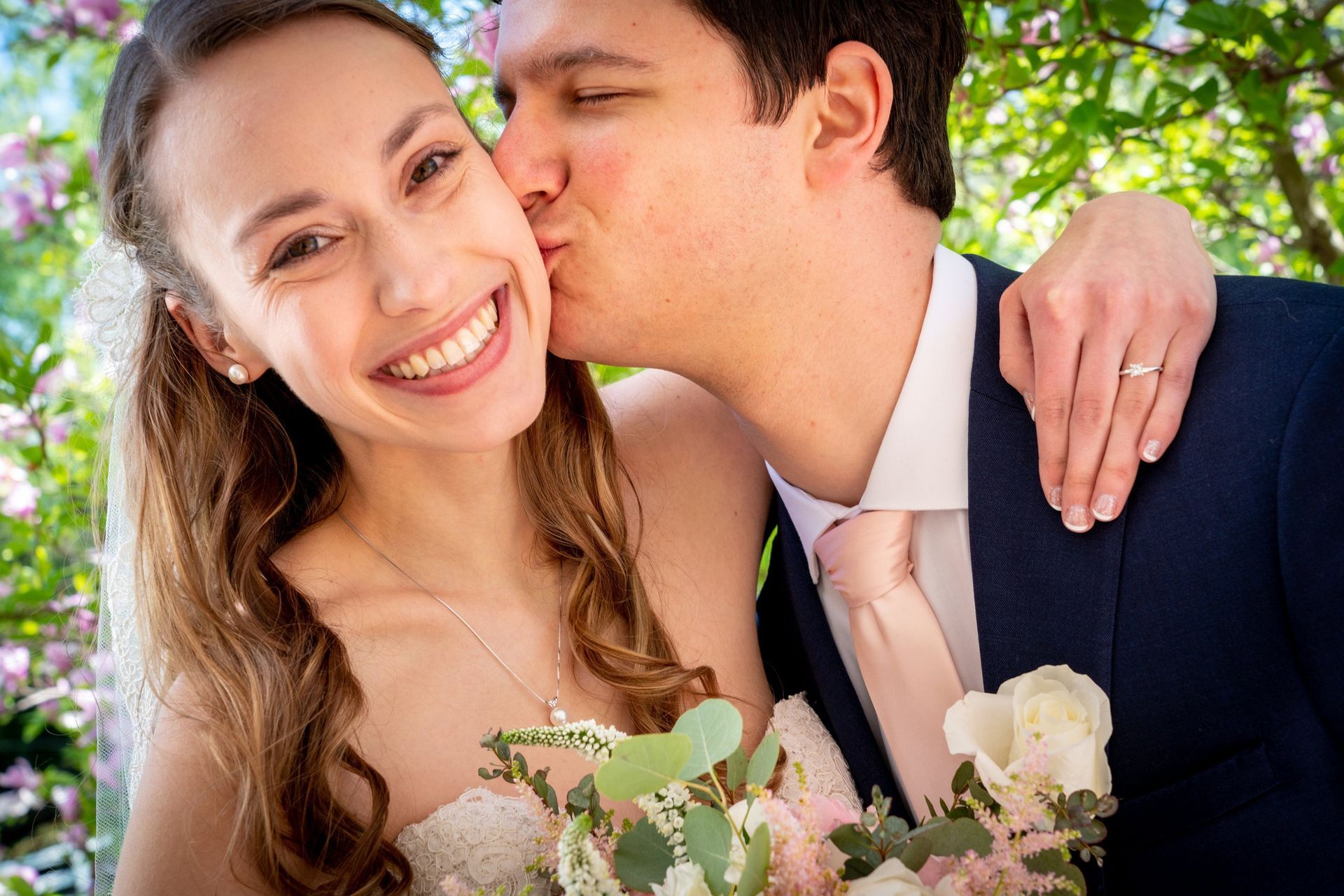 Bride and groom smiling, the groom kissing the bride's cheek. They are in a garden.