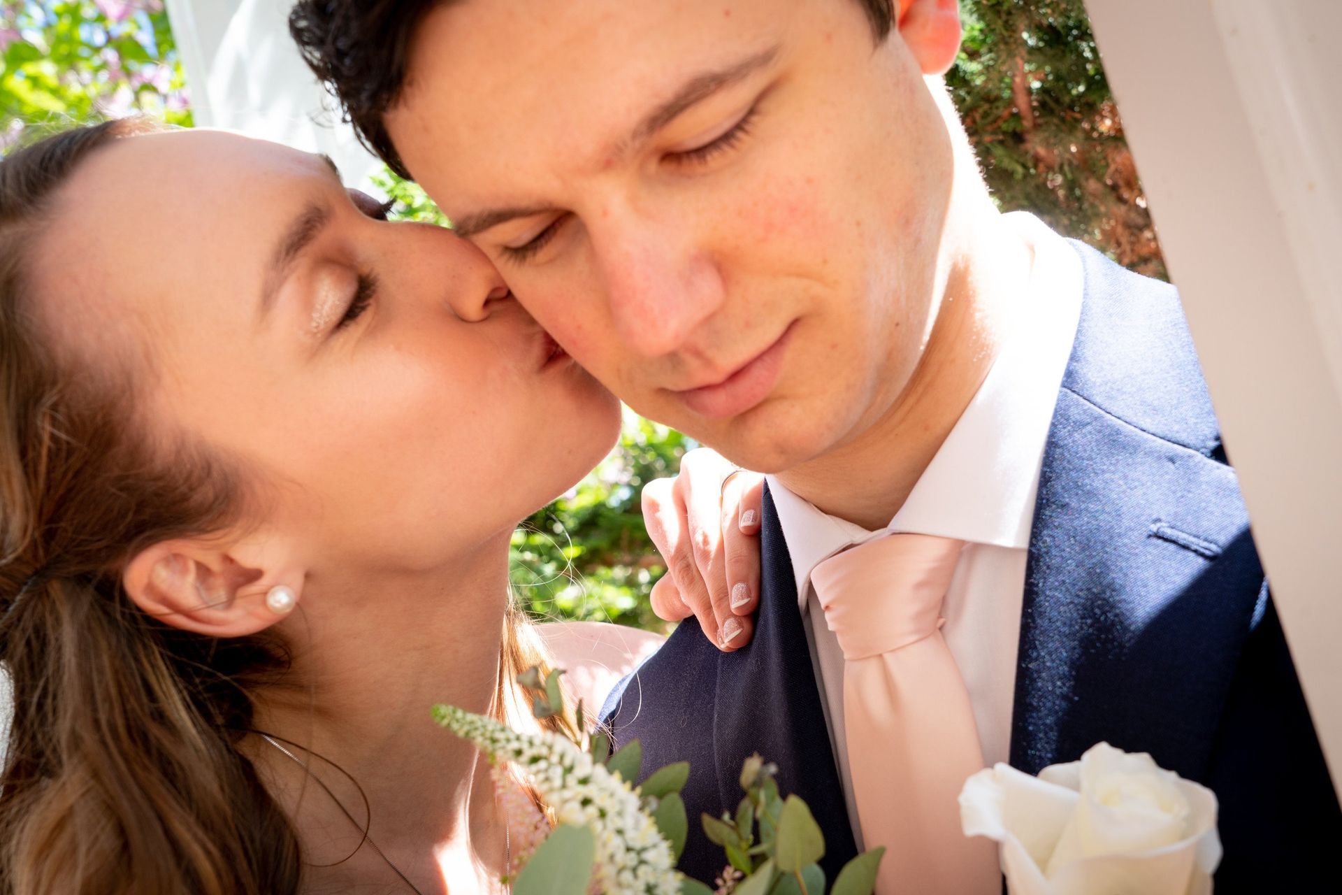 Bride kissing groom's cheek; both have eyes closed. Groom wears navy suit with pink tie. Outdoor setting.