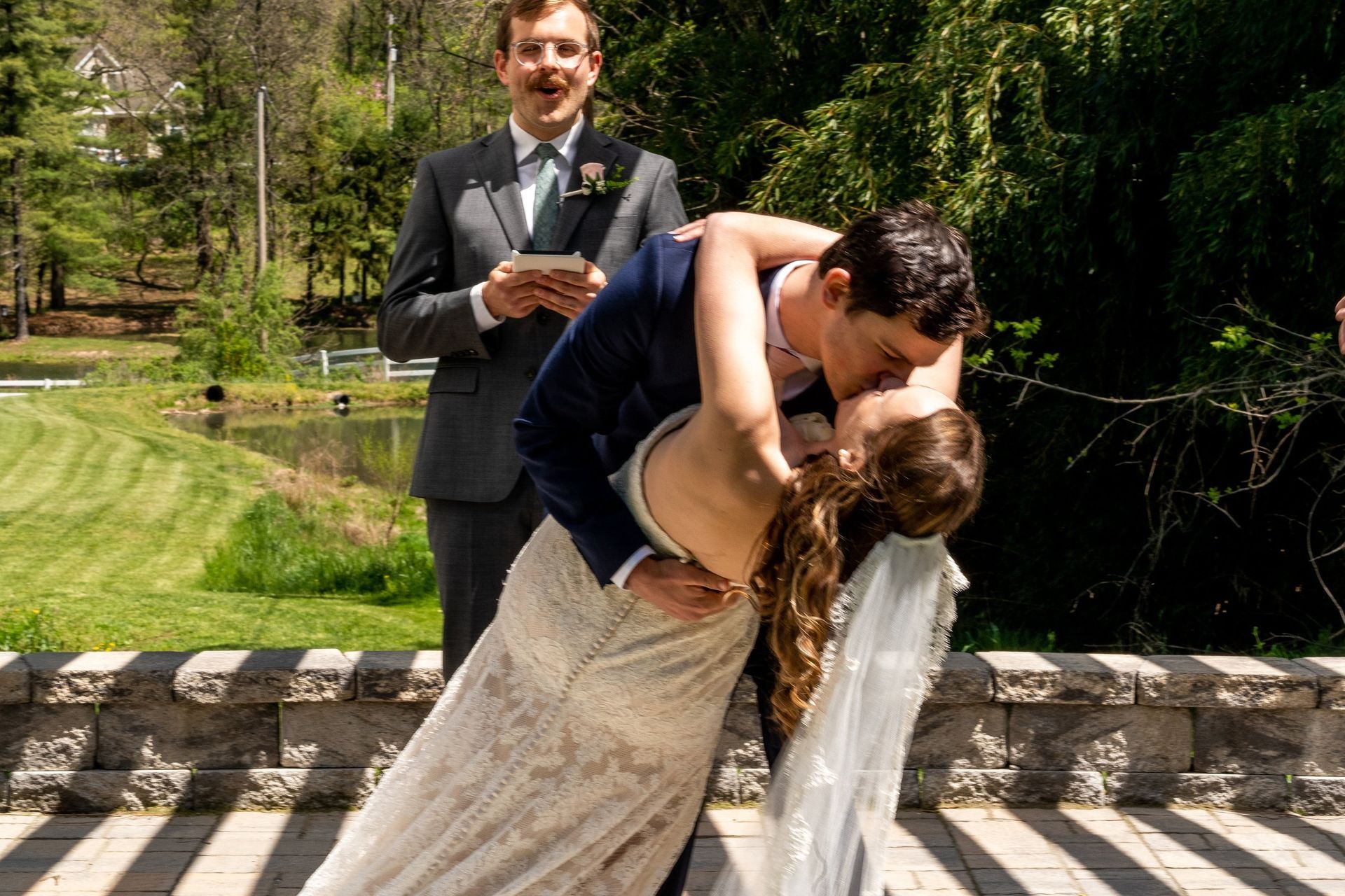 Newlyweds share a kiss during an outdoor ceremony. The officiant smiles, green setting.