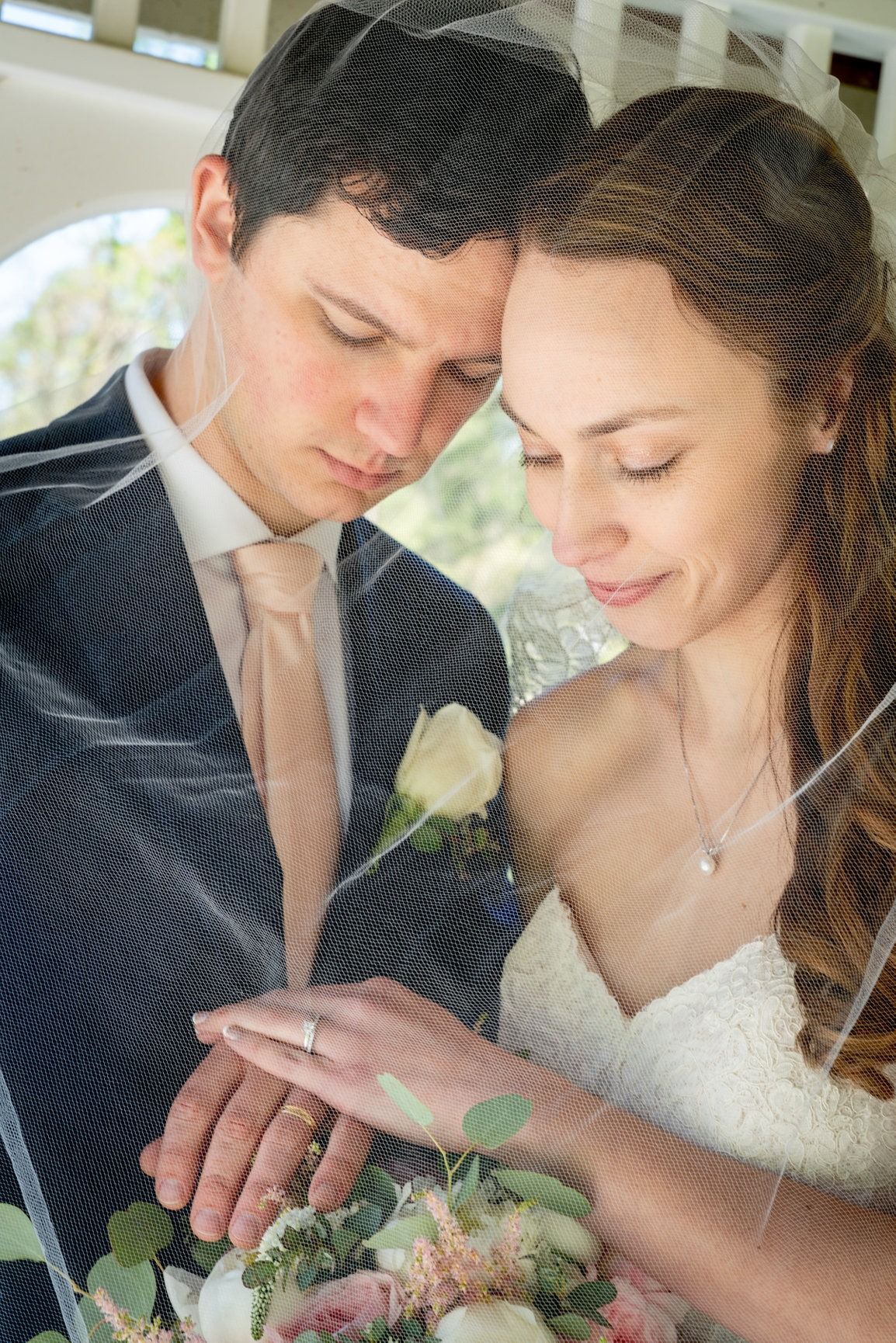 A newlywed couple, veiled, gazes at their hands, a bouquet in the foreground.