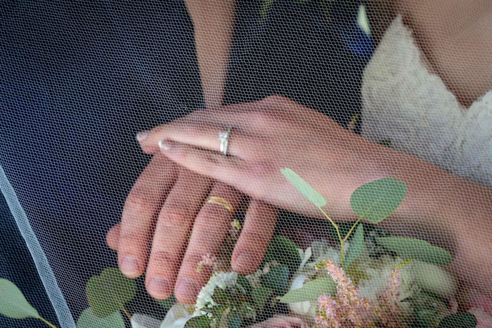 Wedding couple's hands clasped, one with a ring. Veil and bouquet partially visible.