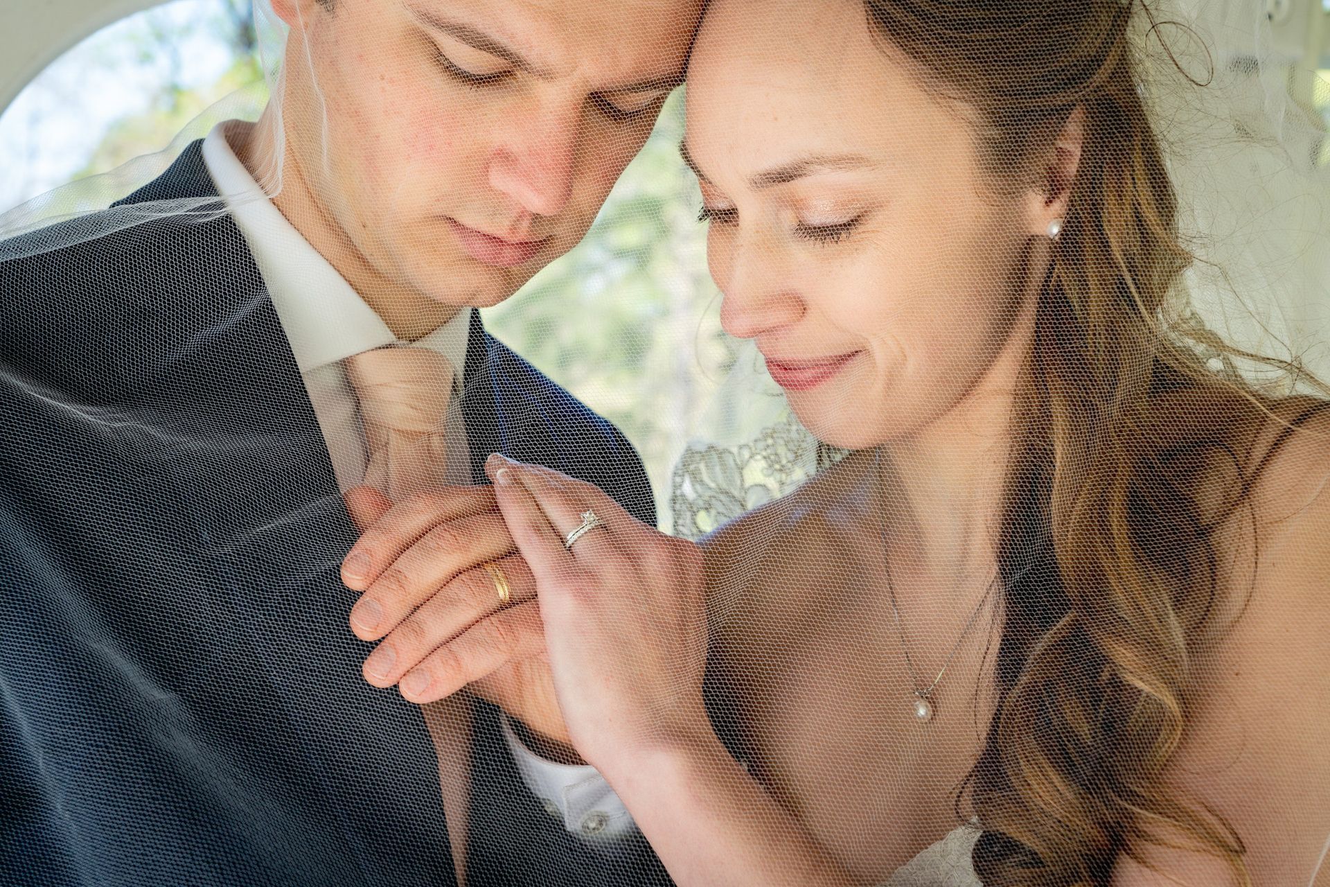 Bride and groom hold hands, tenderly looking down. He wears a navy suit, she wears a veil.