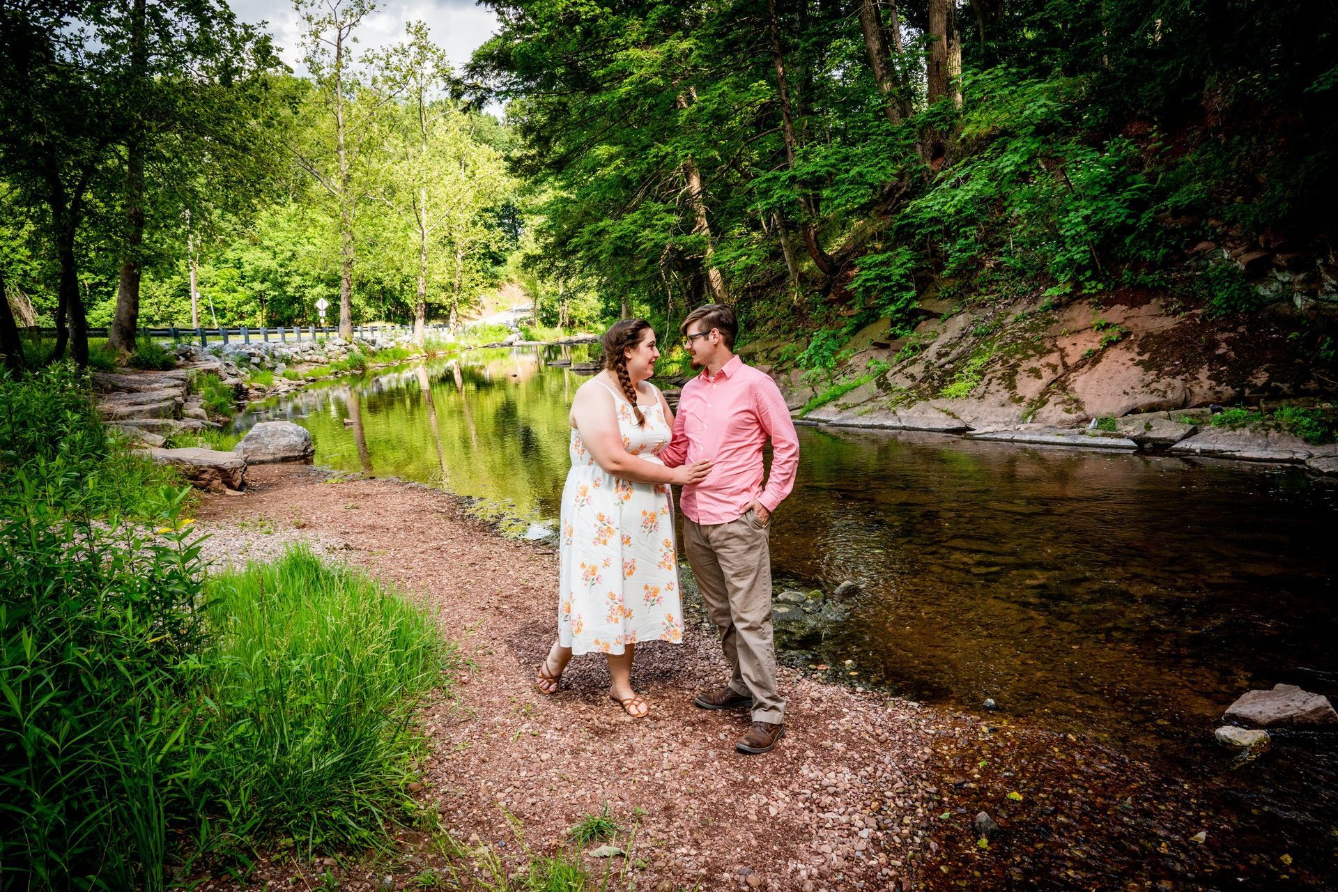 Couple standing by a river, holding hands, looking at each other. Lush green trees surround them.