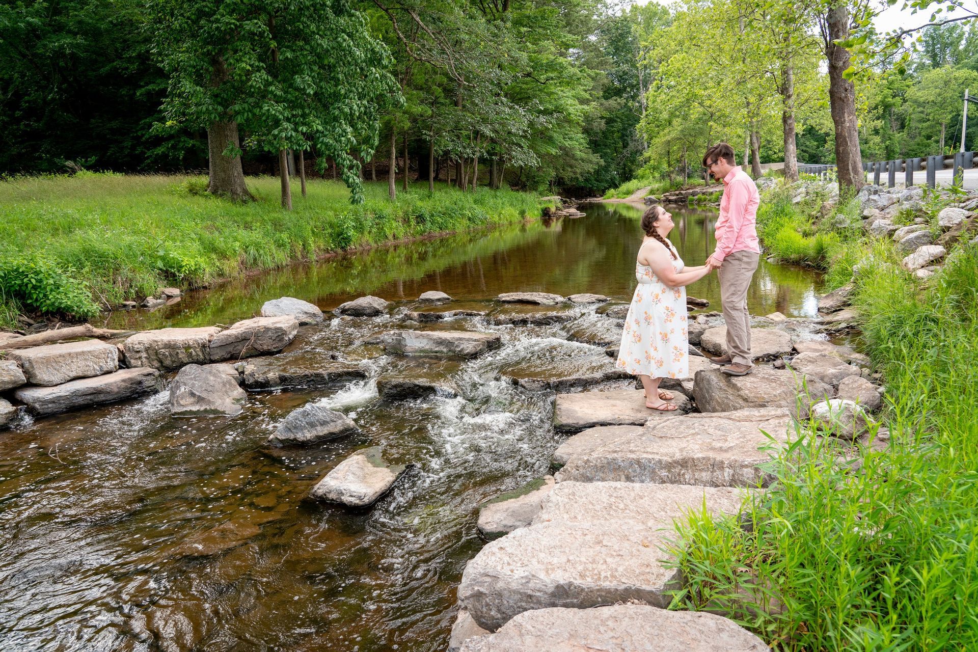 Couple standing on rocks in a stream, holding hands. Woman in floral dress, man in pink shirt, green trees.