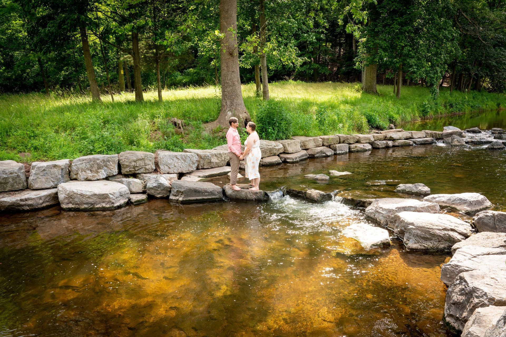 Couple standing hand-in-hand on rocks in a stream; trees and green grass surround them in a park setting.