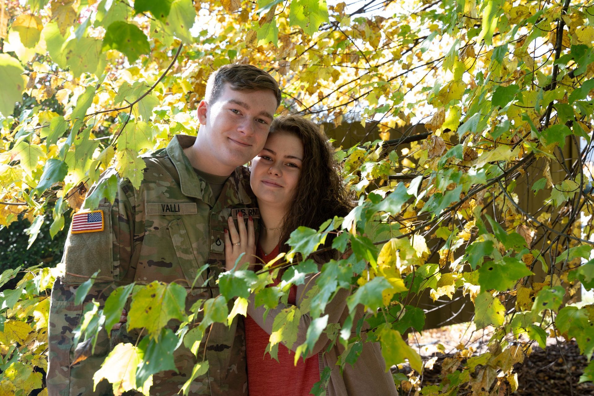 Couple embraces outdoors; man in army uniform, woman in red, framed by fall foliage.
