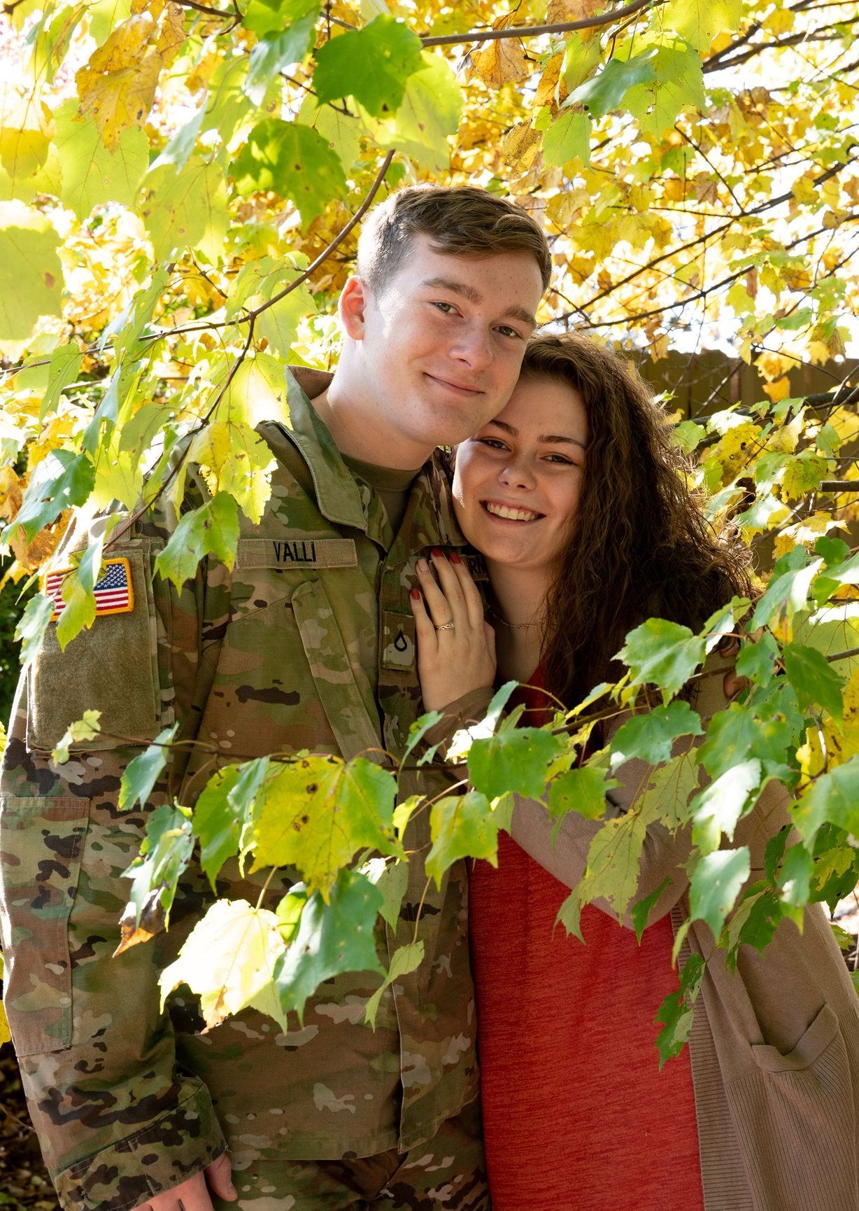 A smiling couple, man in Army uniform, woman in red dress, framed by fall foliage.