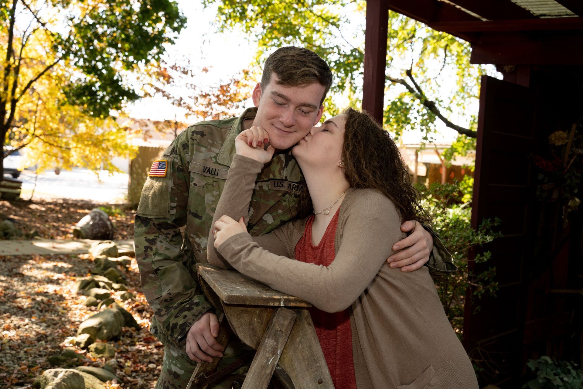 Woman kissing a soldier's cheek; they embrace outdoors, near a rustic wooden structure, with autumn foliage.