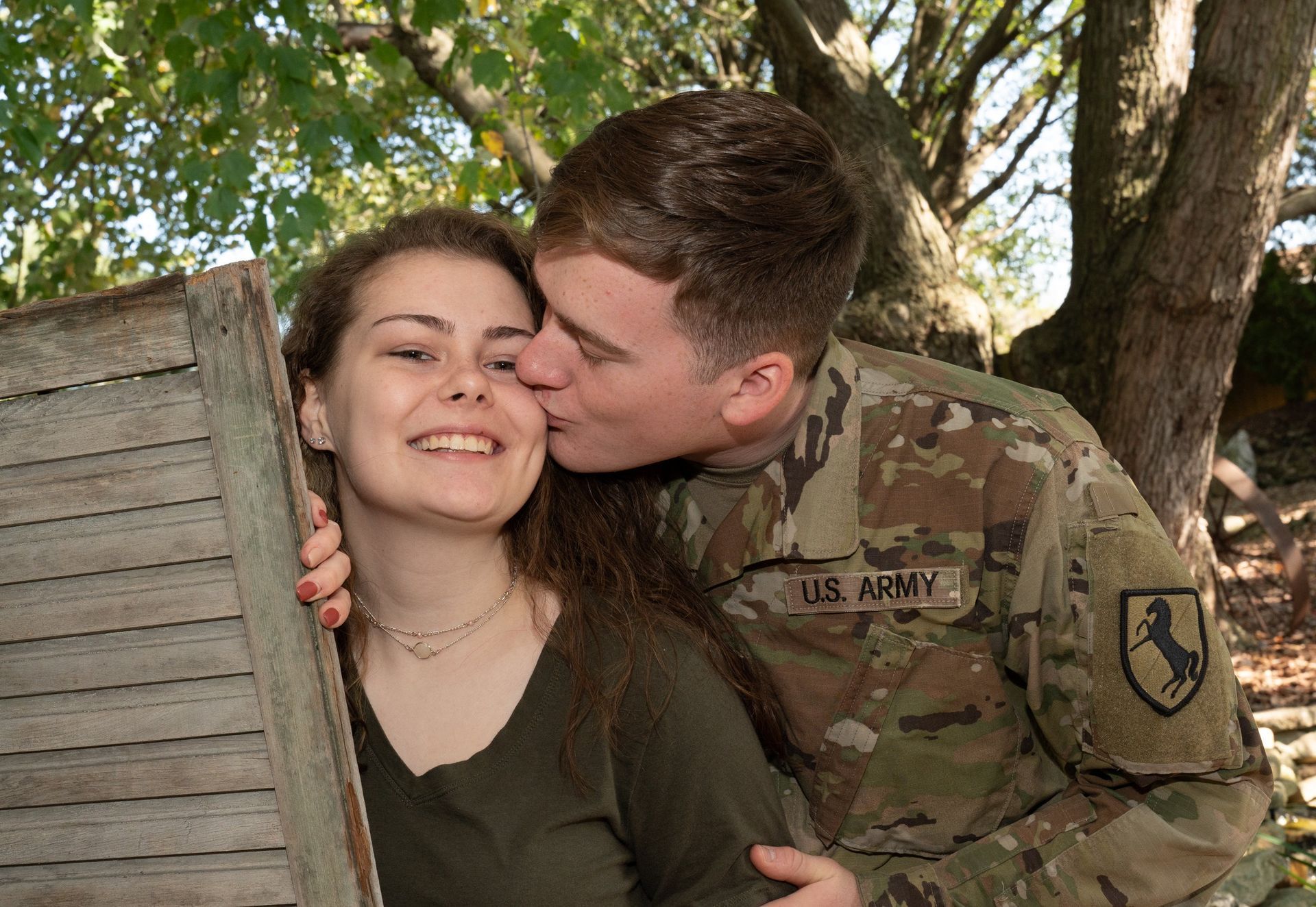 Man in military uniform kisses woman on the cheek; both smile in outdoor setting.