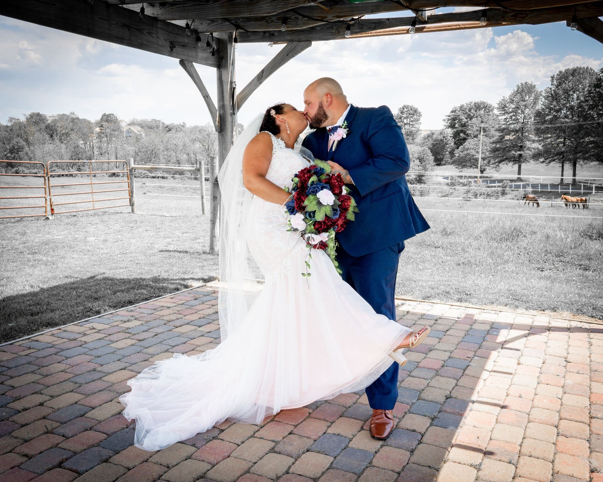 Bride and groom kissing under an outdoor pergola; she's in a white dress, he's in a blue suit.