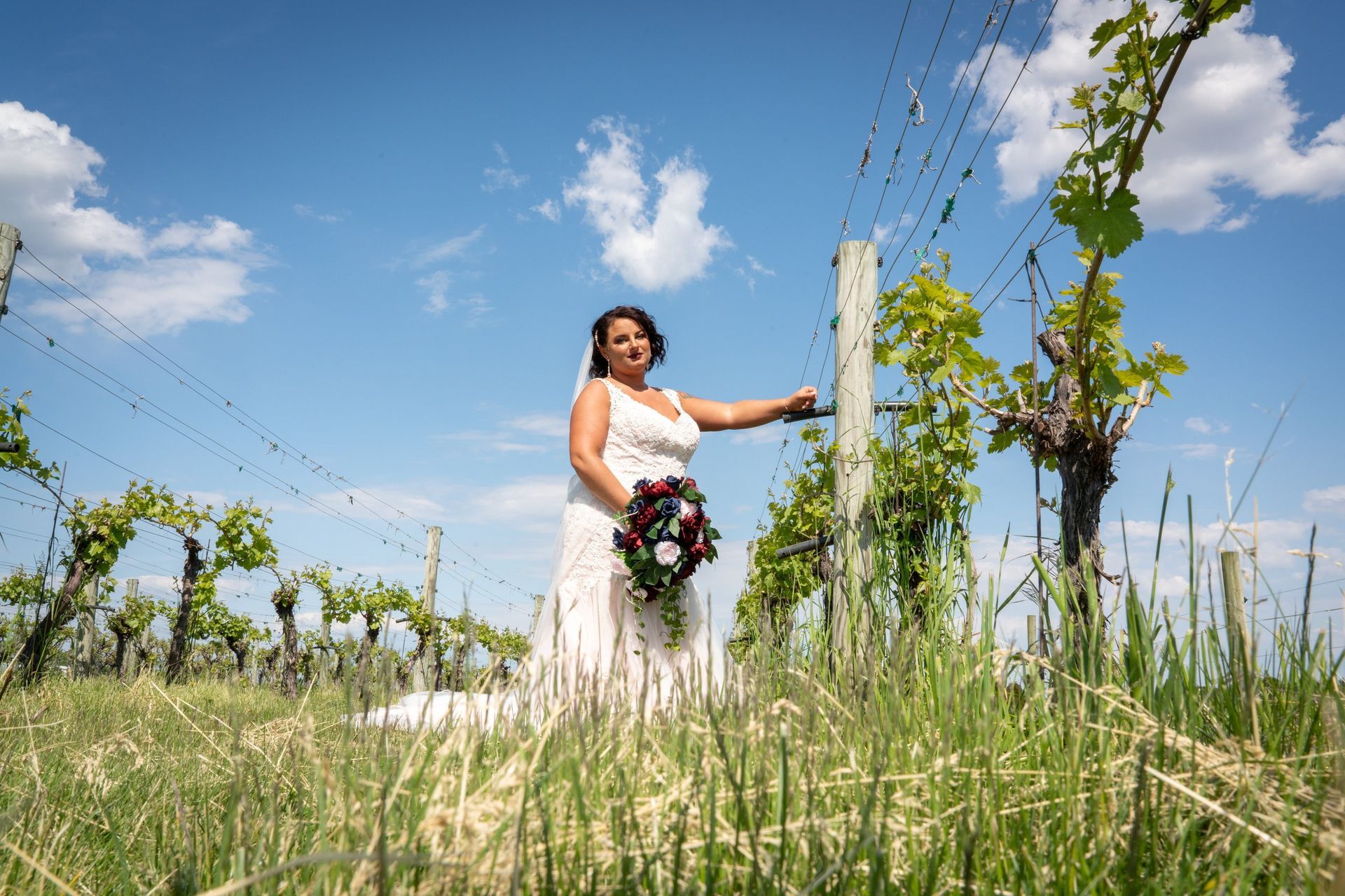 Bride in a white dress poses in a vineyard, holding a bouquet, under a blue sky.