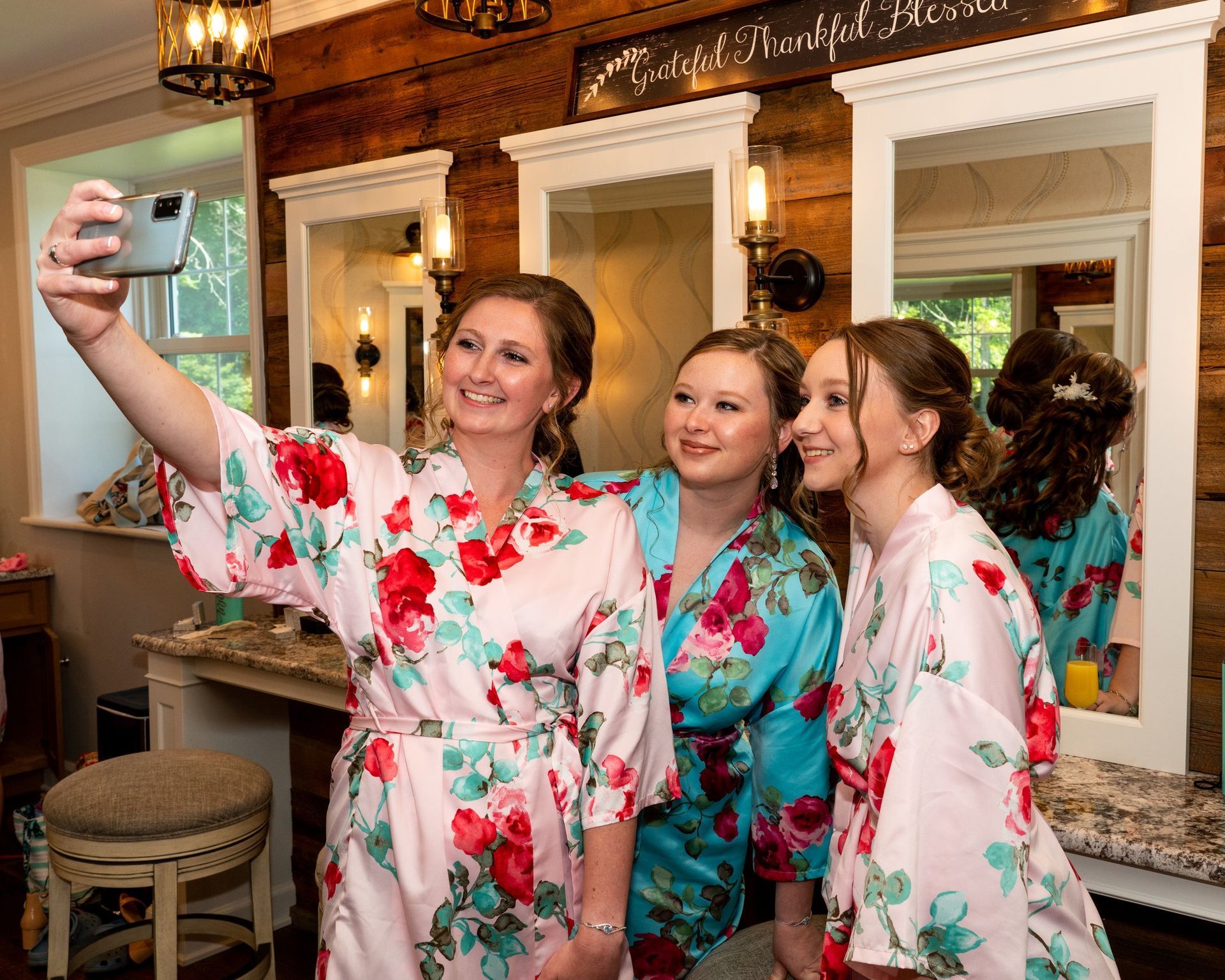 Three women in floral robes take a selfie in front of mirrors; rustic wood background.