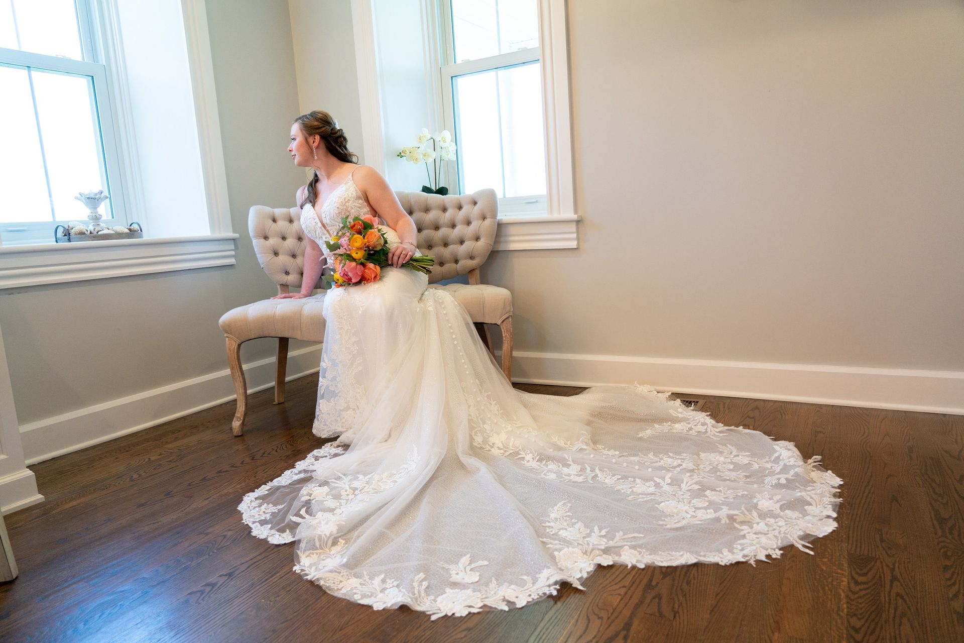 Bride in wedding dress sits on a bench, gazing out a window, holding a bouquet.