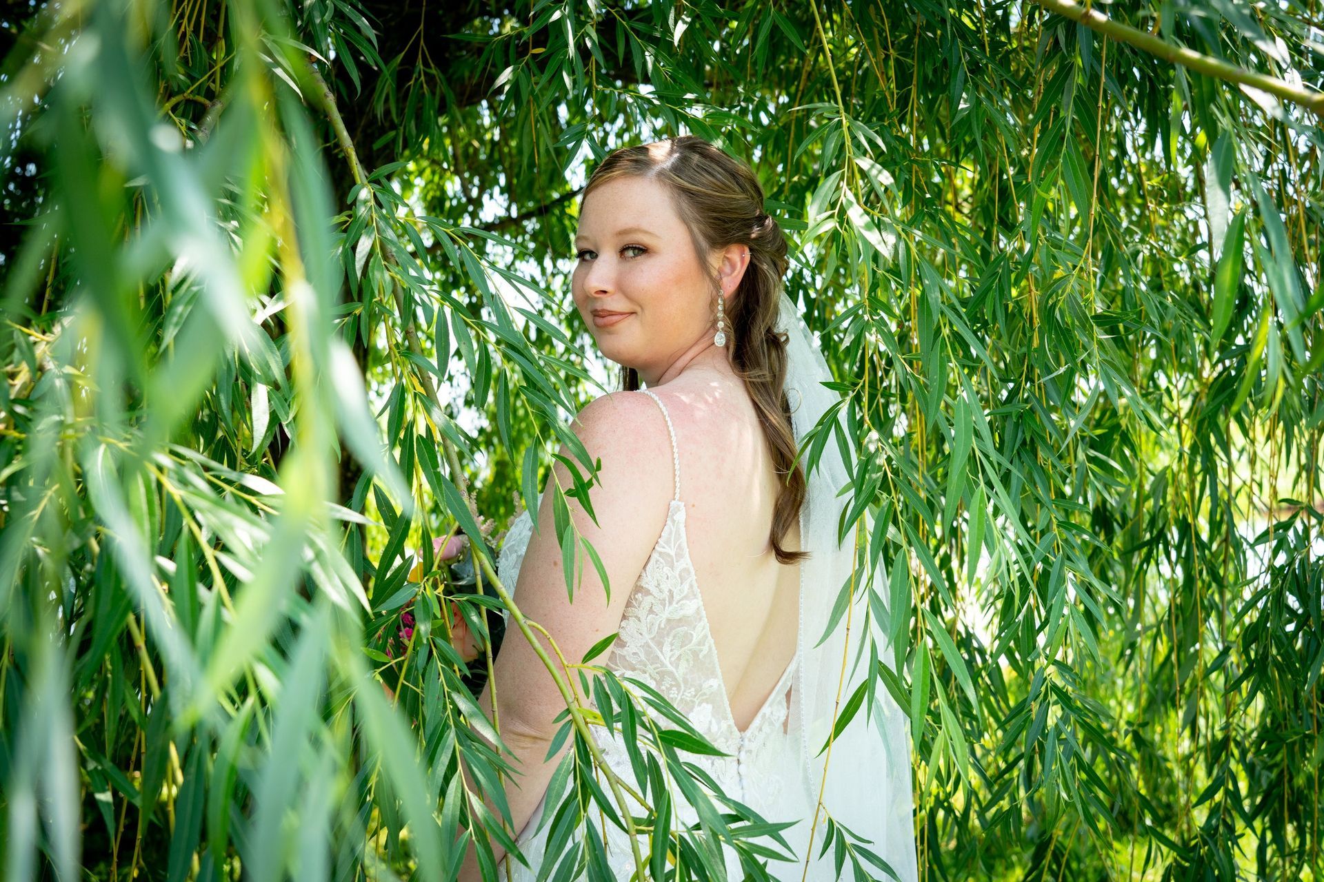 Bride in wedding dress, braided hair, smiling, partially hidden by green willow branches.