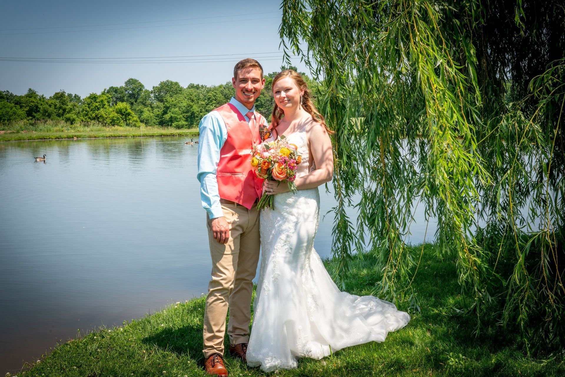 Wedding couple poses by a lake: man in coral vest, woman in white gown, under a weeping willow.