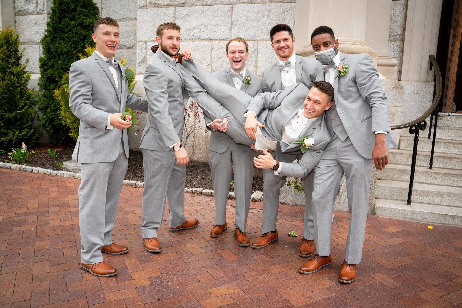 Groomsmen in gray suits lifting and posing with another man; all smiling on brick path.