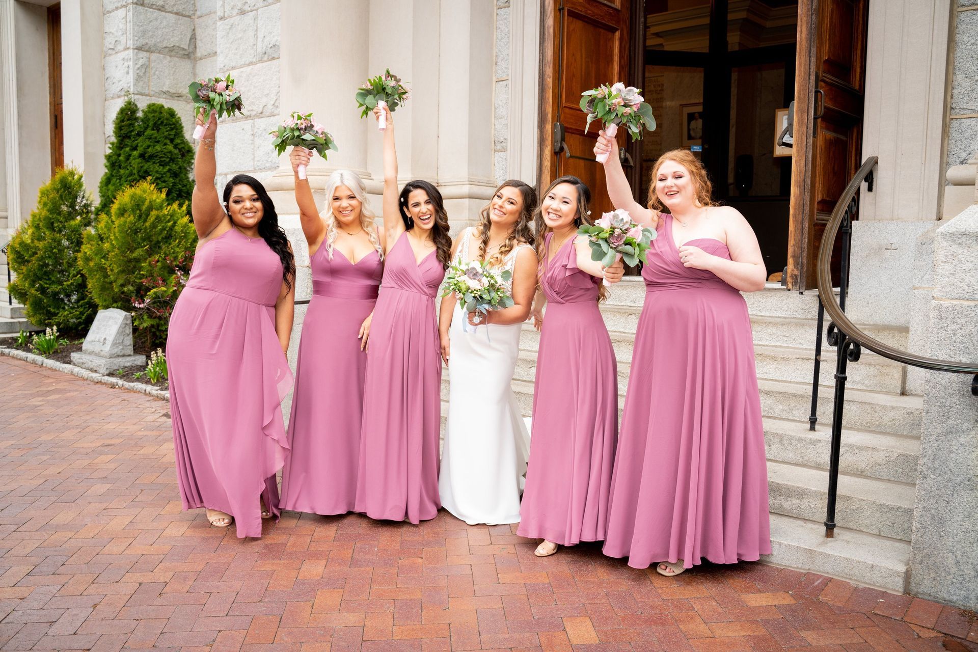 Bridesmaids in mauve dresses and bride in white celebrate outside a church, holding bouquets.
