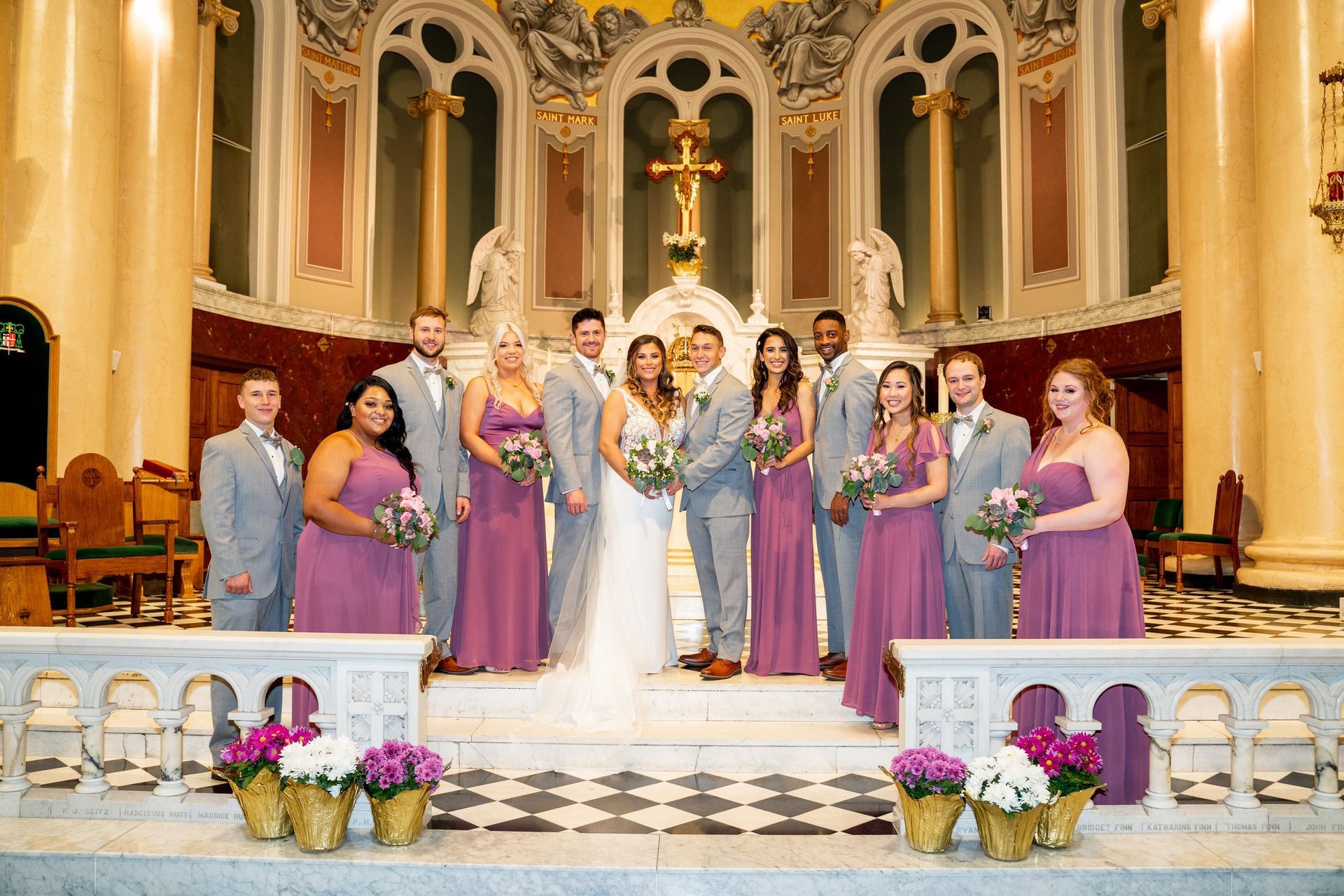 Wedding party posing in a church. Bride and groom centered, surrounded by bridesmaids in purple and groomsmen in gray.