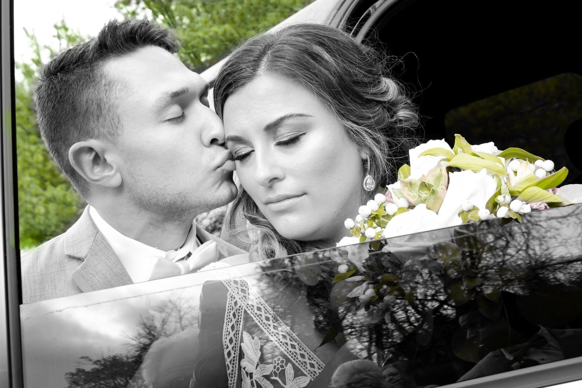 Groom kissing bride's cheek next to a car, holding flowers. Black and white with color accent.
