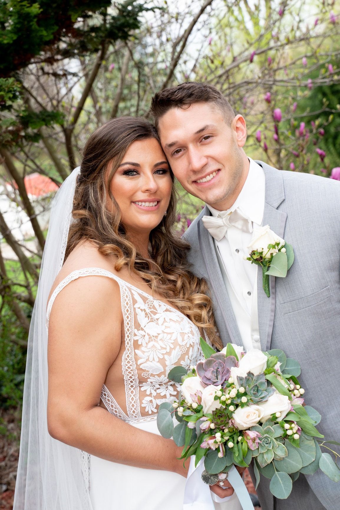 Bride and groom smiling; she holds bouquet, he wears grey suit, outside by trees.