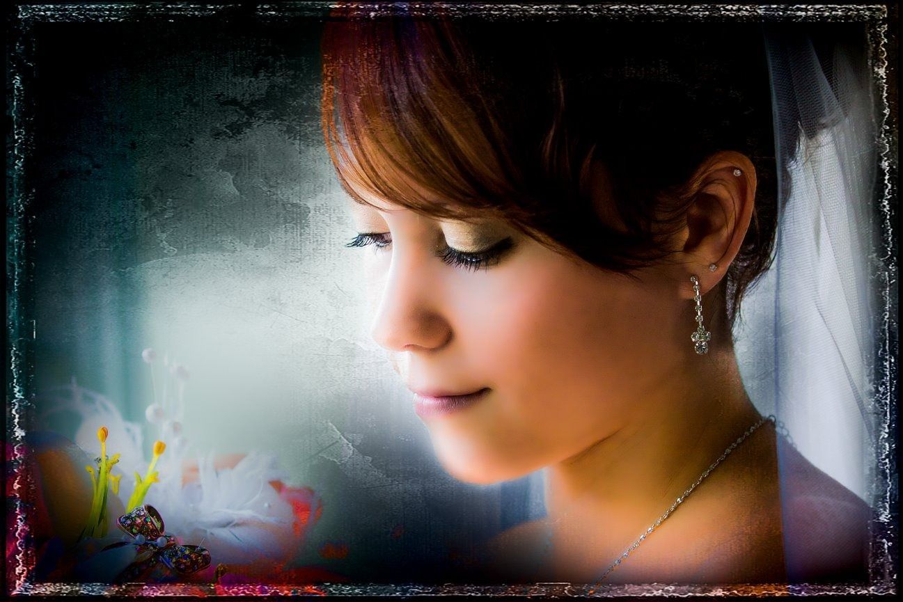 Bride gazes down; brown hair, veil, and jewelry; colorful flowers at left; window background.