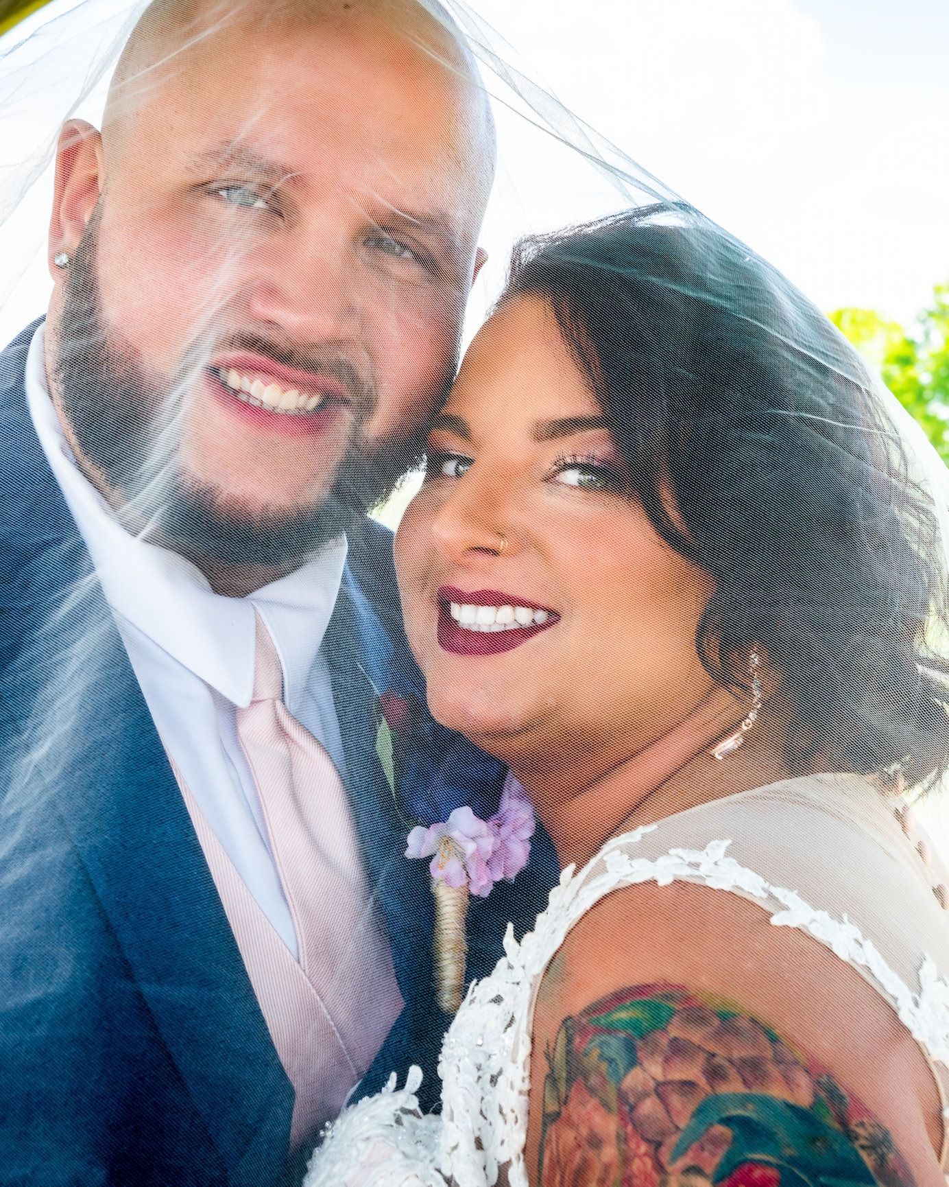 Newlywed couple smiling under a veil; the woman with a shoulder tattoo, wearing a white dress and the man a suit.
