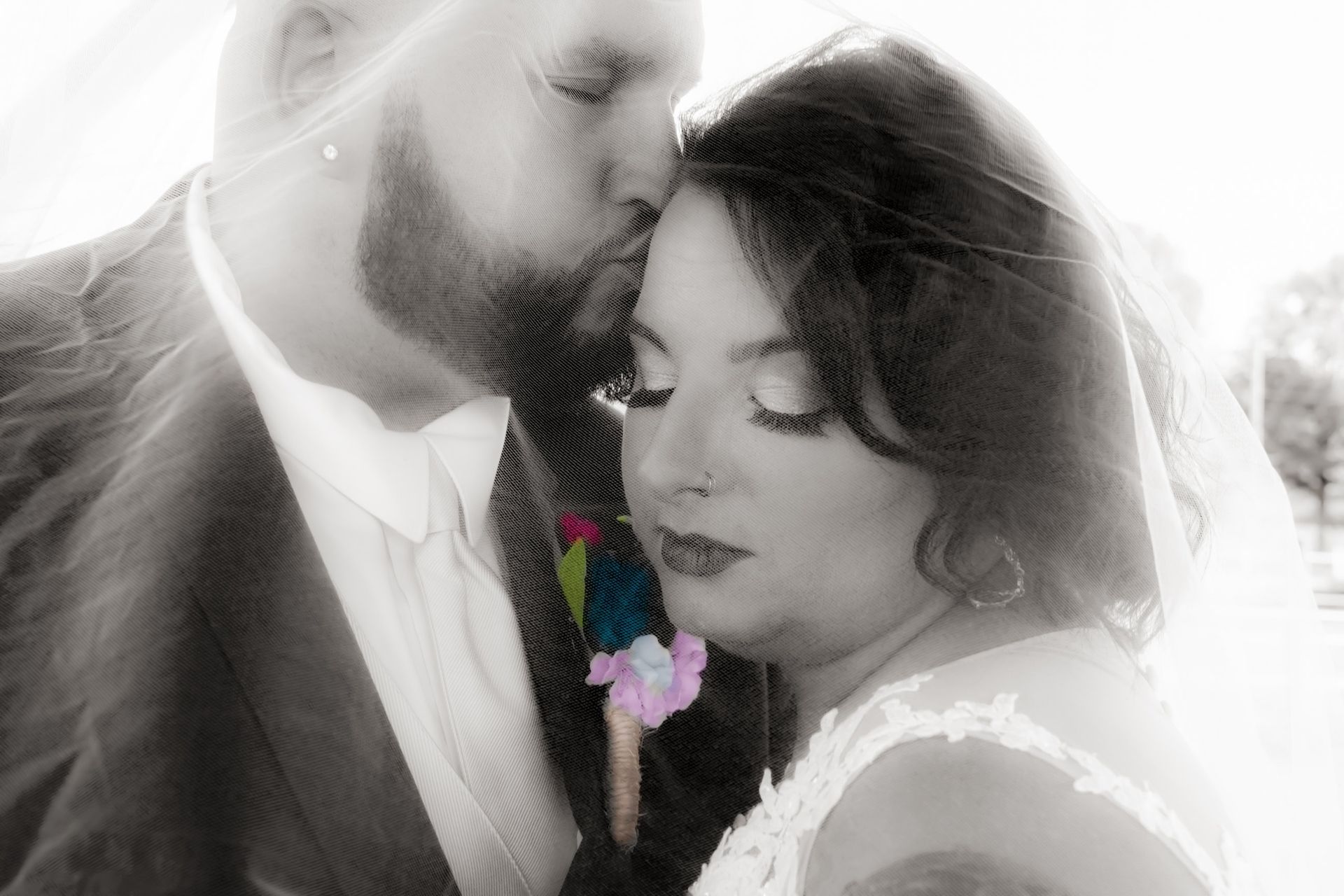 Groom kisses bride's forehead. Black and white photo, outdoor setting, couple close, flower on suit.