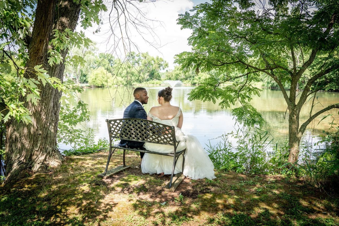 Newlyweds sit on a bench by a lake, framed by trees. The bride wears a white gown, the groom a suit.