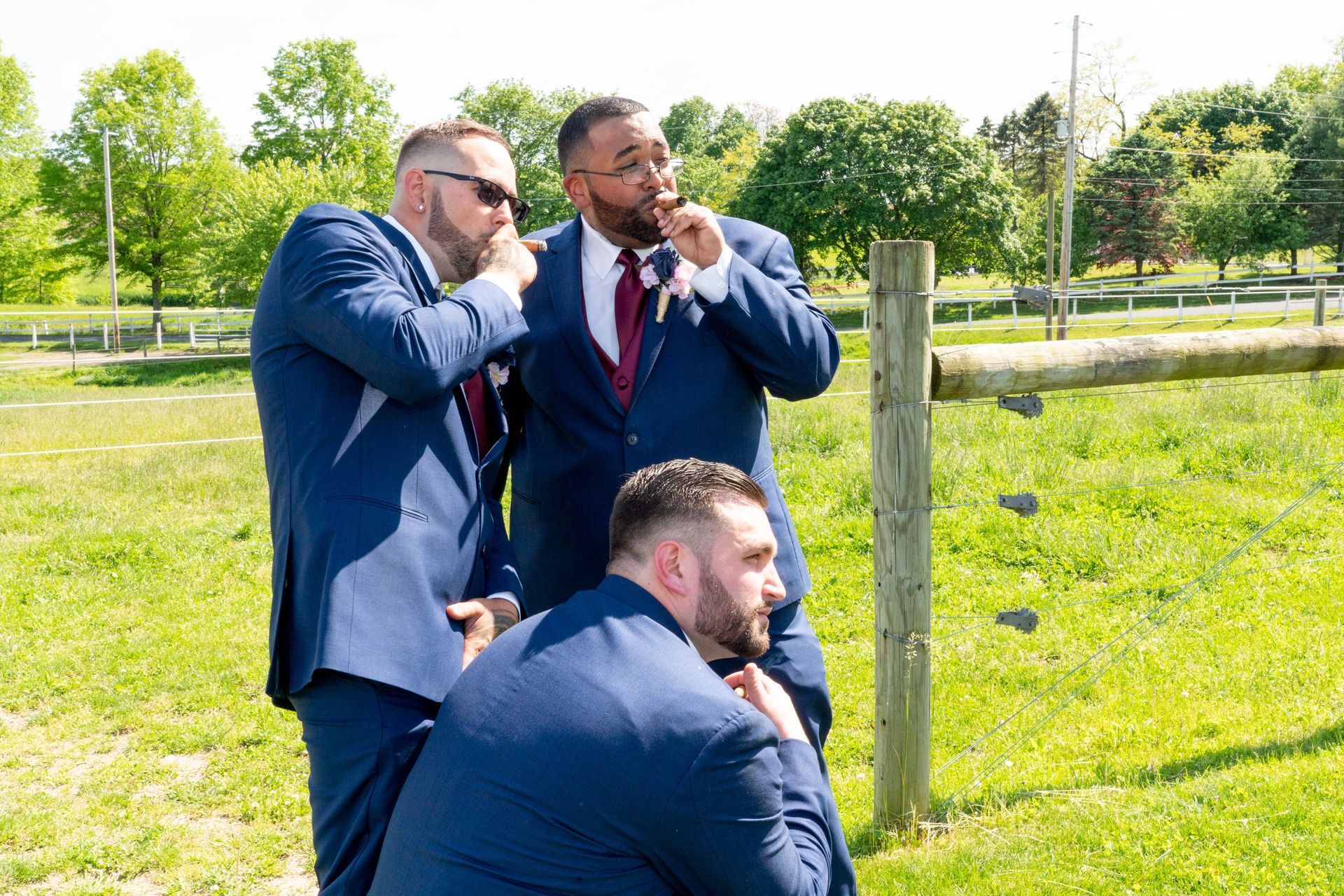 Three men in blue suits, one smoking, near a wooden fence in a sunny outdoor field.