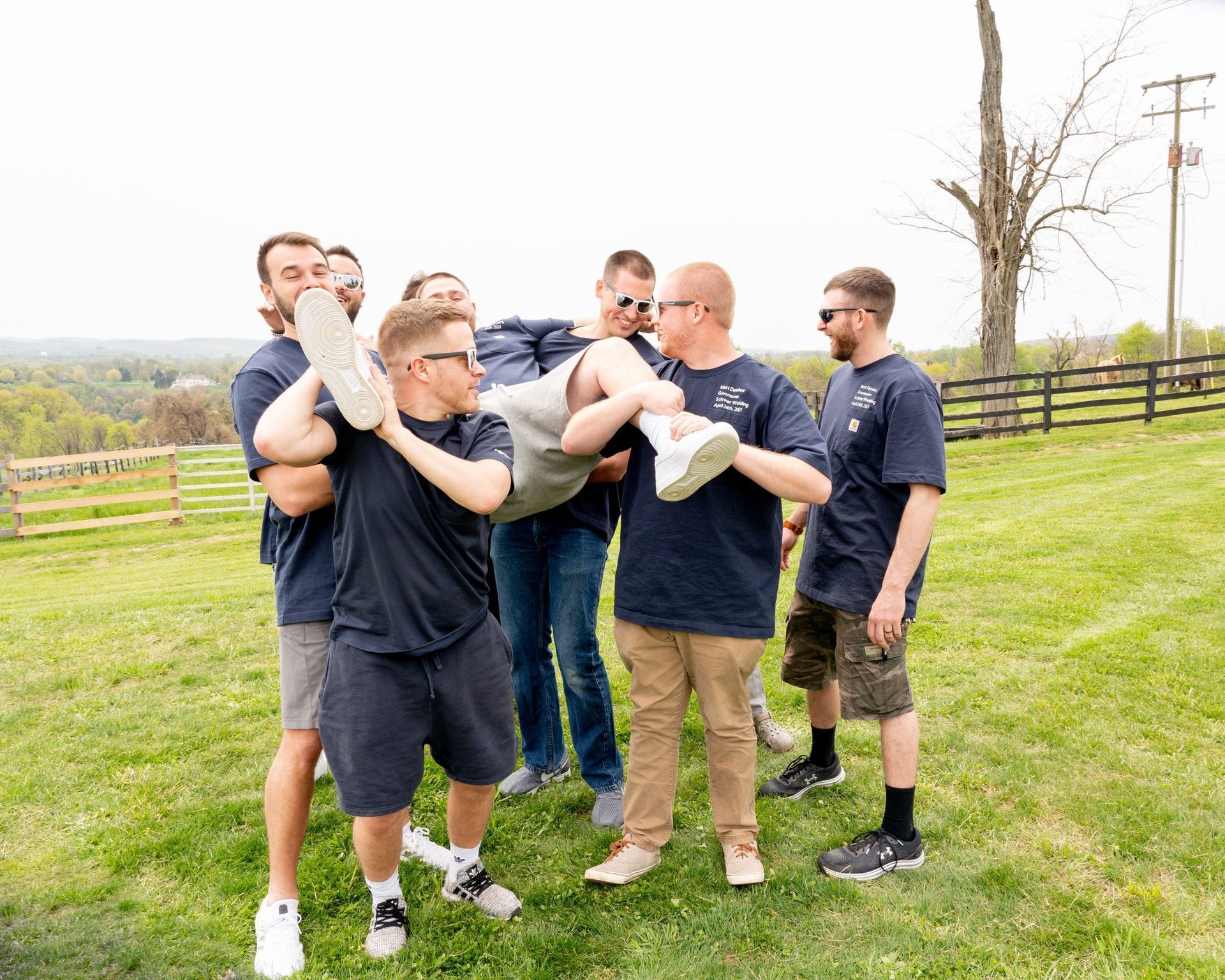 Men carrying a person outdoors on a sunny day.  Group is wearing blue shirts and casual clothes in a grassy field.