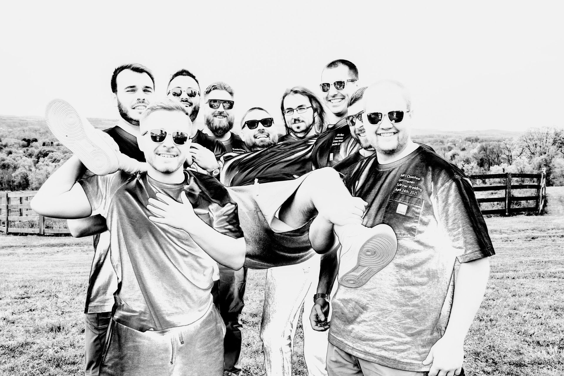 Group of men outside, some holding others up, wearing sunglasses, in field.