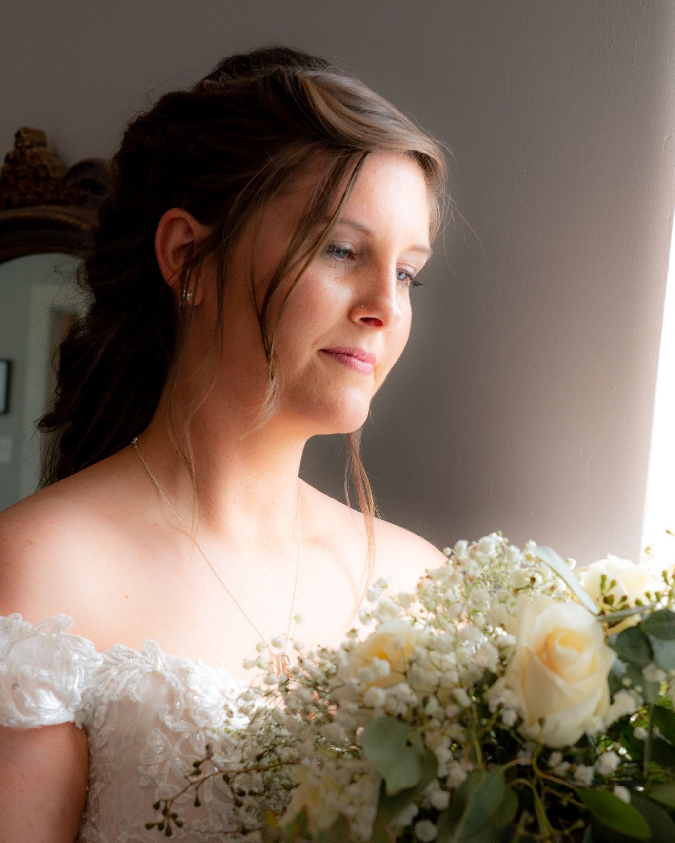 Bride holding flowers, looking thoughtful by window. Wearing white, off-the-shoulder dress, with bouquet of white roses.