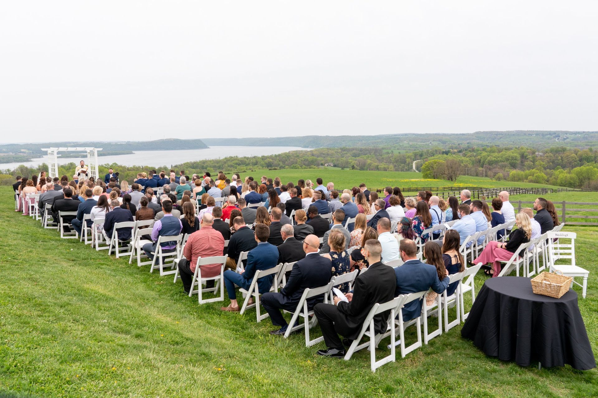Wedding ceremony on a grassy hill with a view of a lake and trees. Guests seated in rows, a ceremony arch.