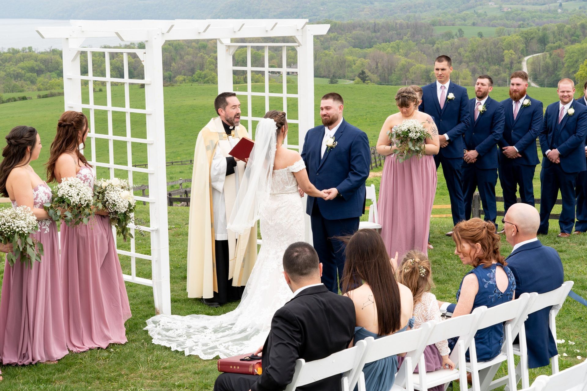 Wedding ceremony outdoors with bride and groom, officiant, wedding party, and guests.