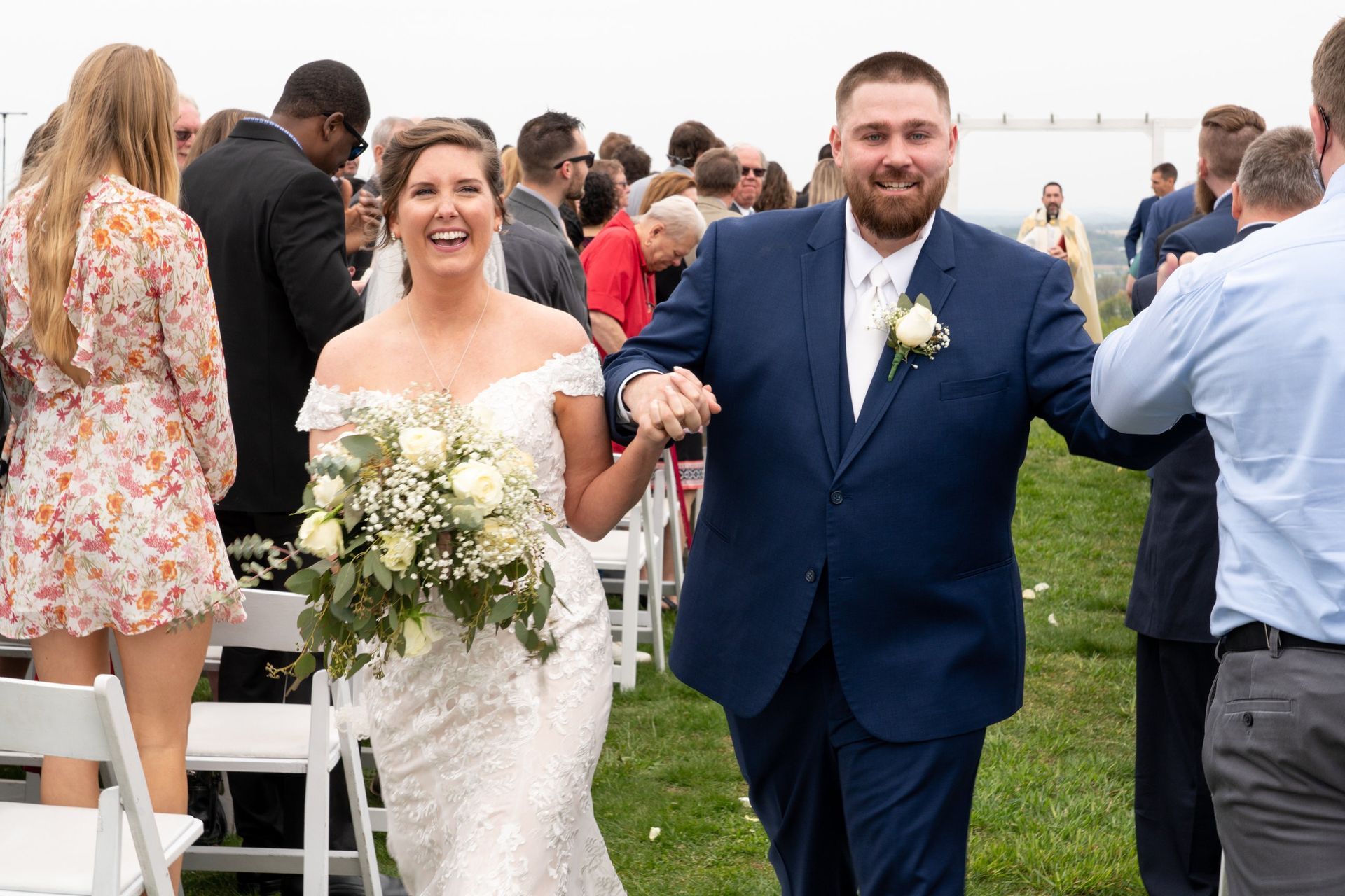 Newly married couple walking down aisle, smiling. Bride in white dress, groom in blue suit. Outdoor ceremony.