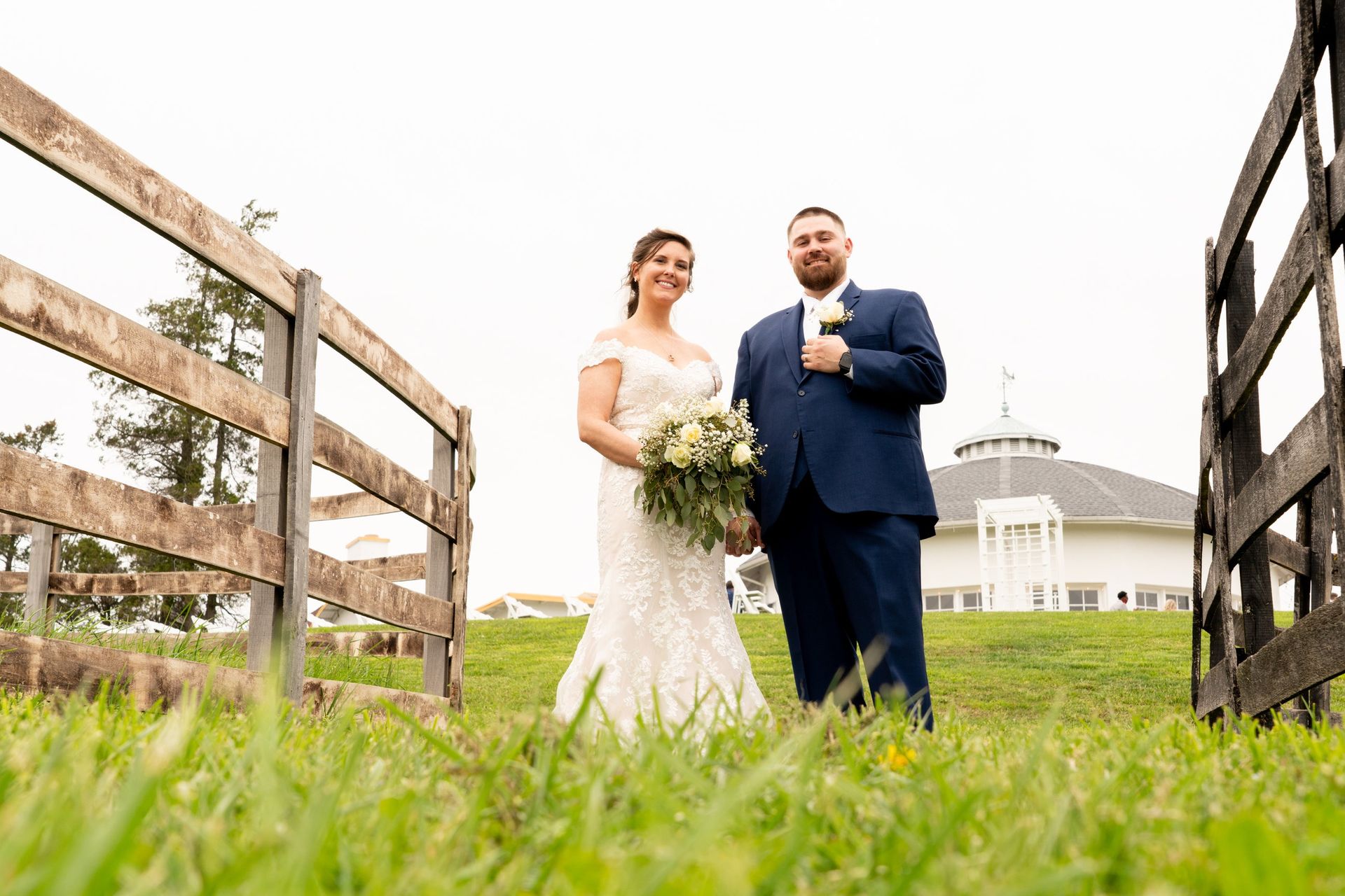 Newlyweds pose, holding hands, framed by a wooden fence, with a building in the background.