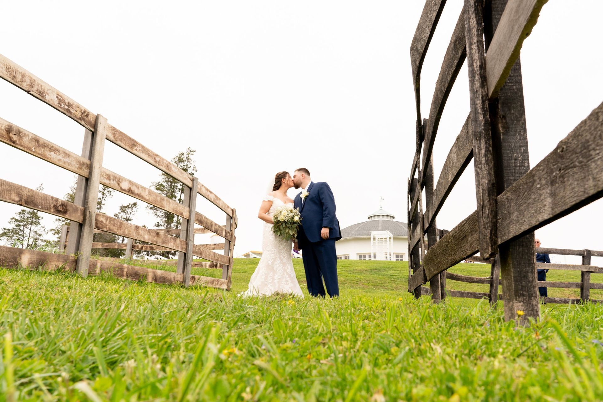 Bride and groom kissing outdoors between wooden fences, wearing wedding attire, green grass, white building in background.