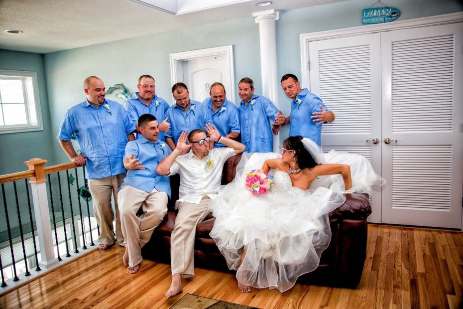 Wedding party photo: Bride in gown and groom making silly faces, groomsmen in blue shirts, beach house setting.