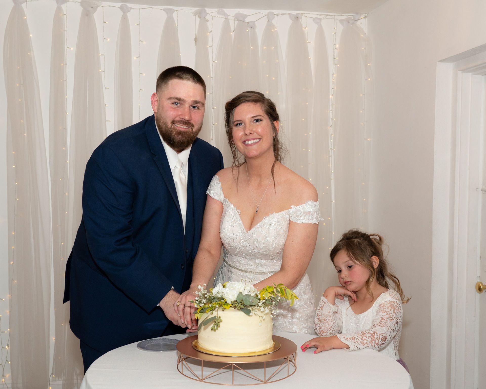 Wedding couple cutting cake, girl watches. Navy suit, white dress, smiles, indoor, cake stand.