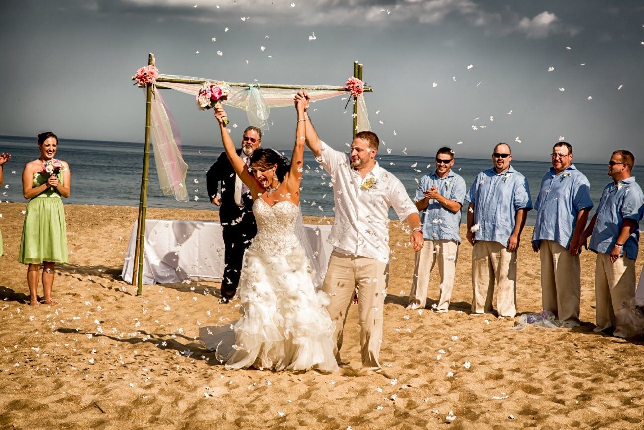 Bride and groom on beach, holding hands up under a bamboo arch, confetti falling. Groomsmen and bridesmaid celebrate.