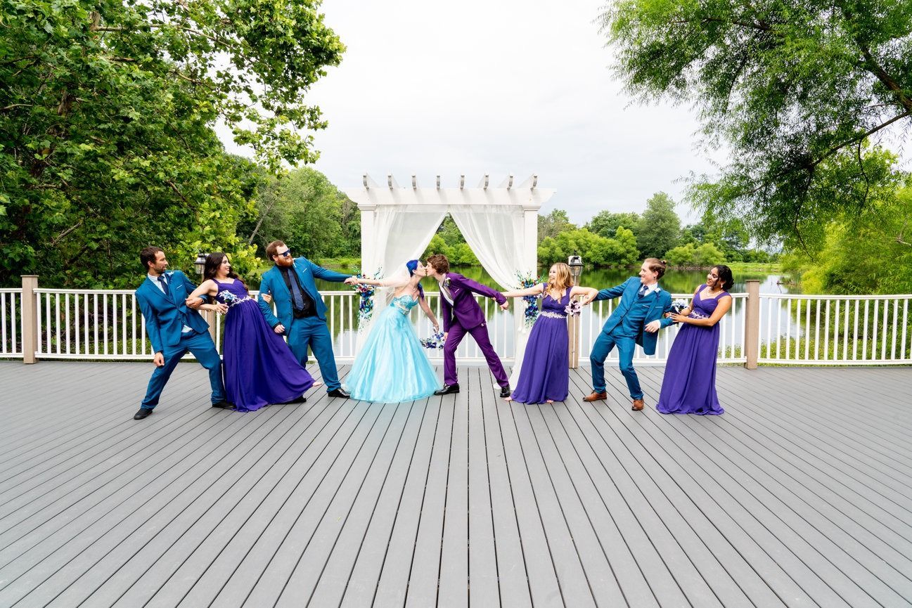 Wedding party poses on a deck: groomsmen in blue pull bride and groom in purple, against a gazebo and lake.